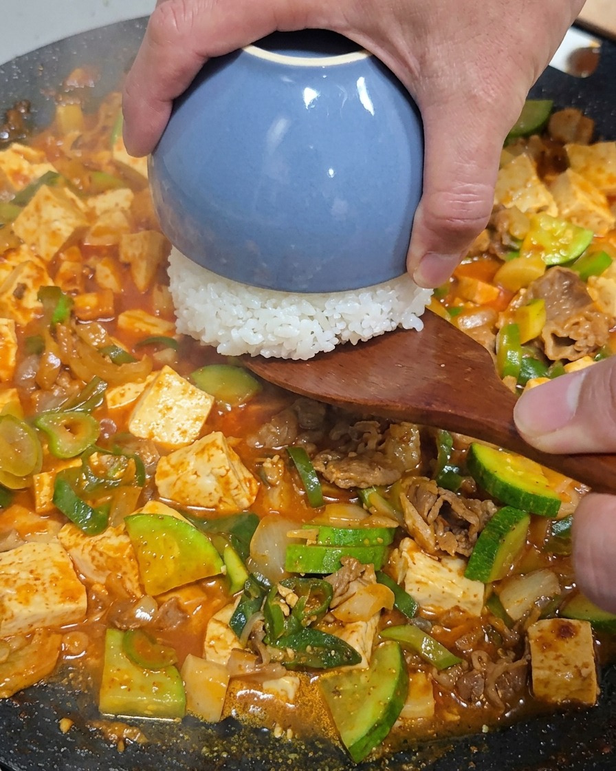 Mano usando una espátula de madera para colocar un pequeño cuenco azul con arroz blanco boca abajo en el centro de la sartén con el estofado.