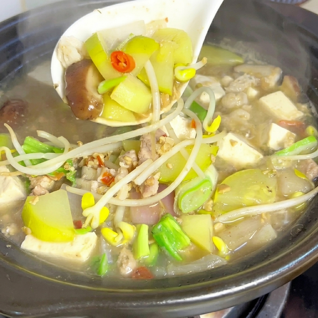 Un cucharón blanco levantando suavemente una mezcla de verduras, tofu y carne de una humeante y burbujeante olla de barro con estofado de pasta de soja.