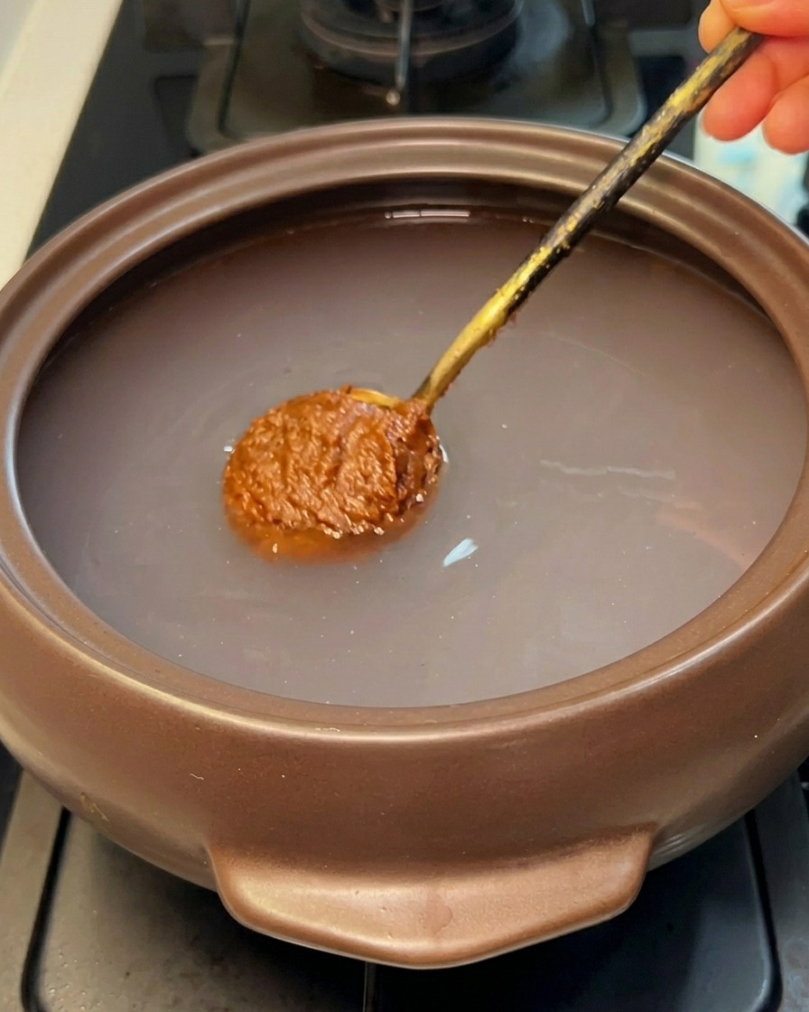 Una cucharada de pasta de soja coreana marrón sumergiéndose en el agua clara de una olla de piedra.