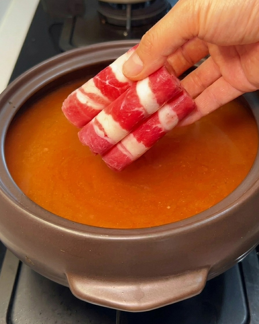 Rollitos de carne de res en rodajas finas siendo añadidos al caldo rojo a fuego lento.