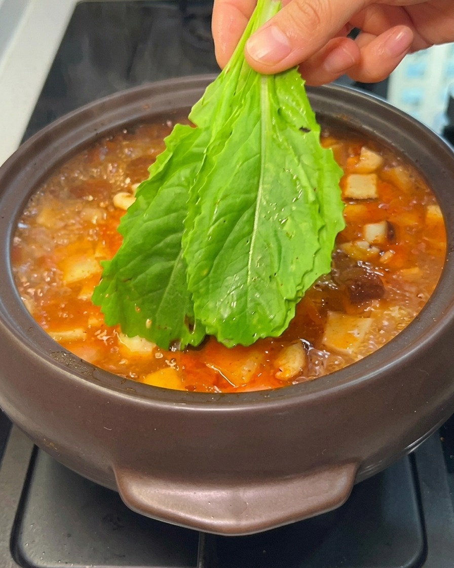 Una mano introduciendo verduras de hoja verde frescas en un estofado coreano hirviendo.