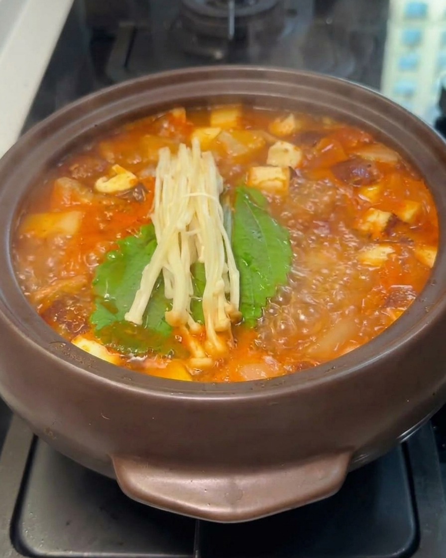 Un manojo de champiñones enoki frescos descansando sobre hojas verdes dentro de una olla hirviendo de estofado rojo.