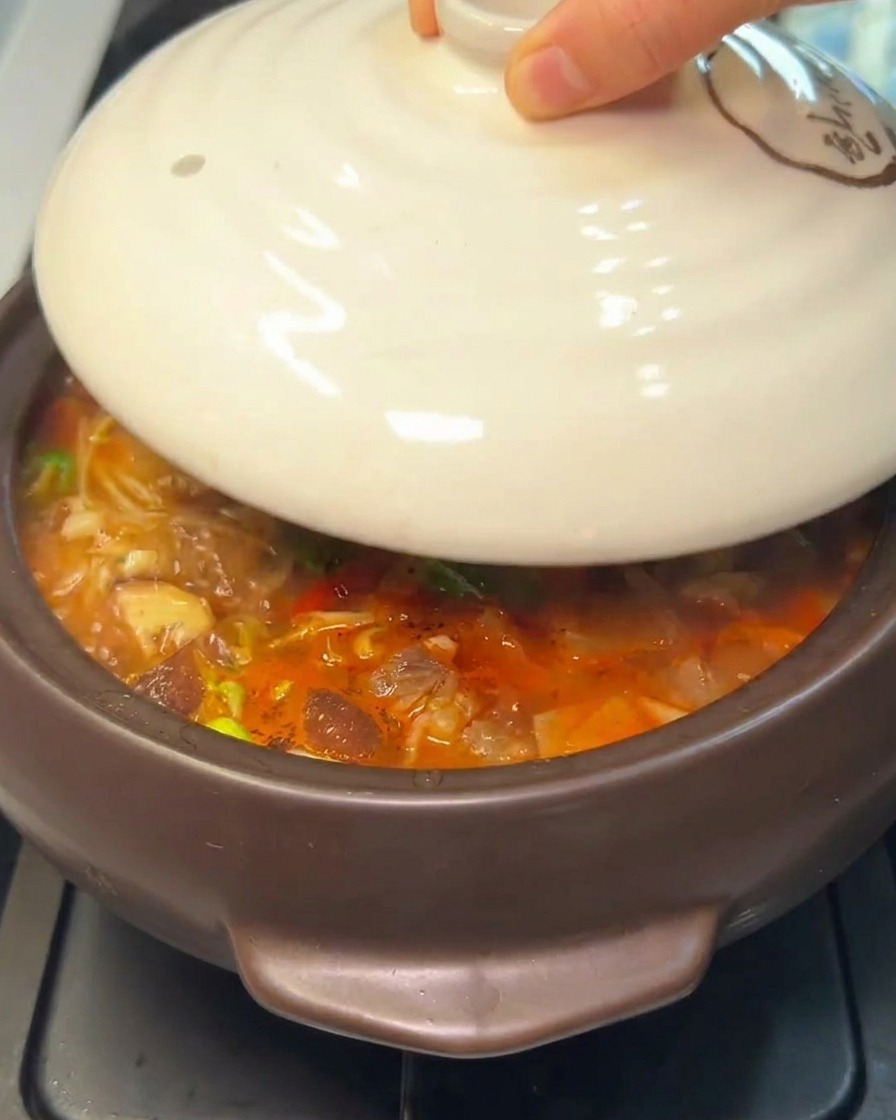 Una mano colocando una tapa de cerámica blanca sobre una humeante olla de piedra con estofado coreano.