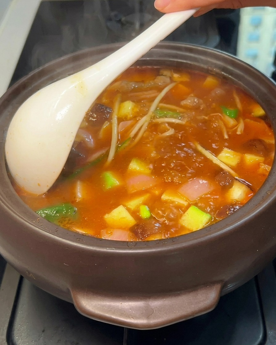 Un primer plano de un estofado coreano hirviendo vigorosamente en una olla oscura, siendo removido con un cucharón blanco para revelar tofu, calabacín, champiñones y carne en un caldo opaco de color rojo anaranjado.