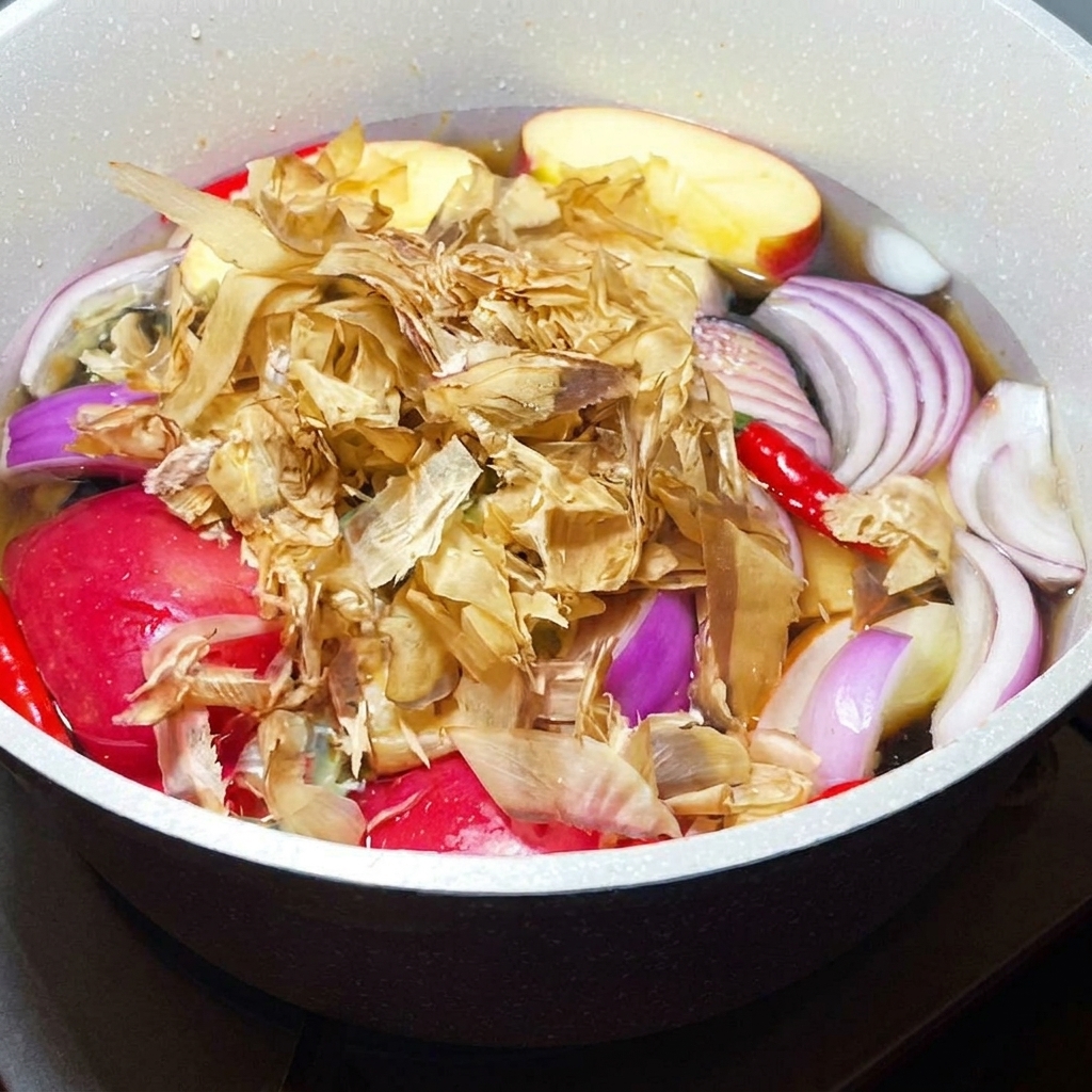 Copos de bonito seco añadiéndose a una olla a fuego lento llena de verduras y frutas aromáticas.