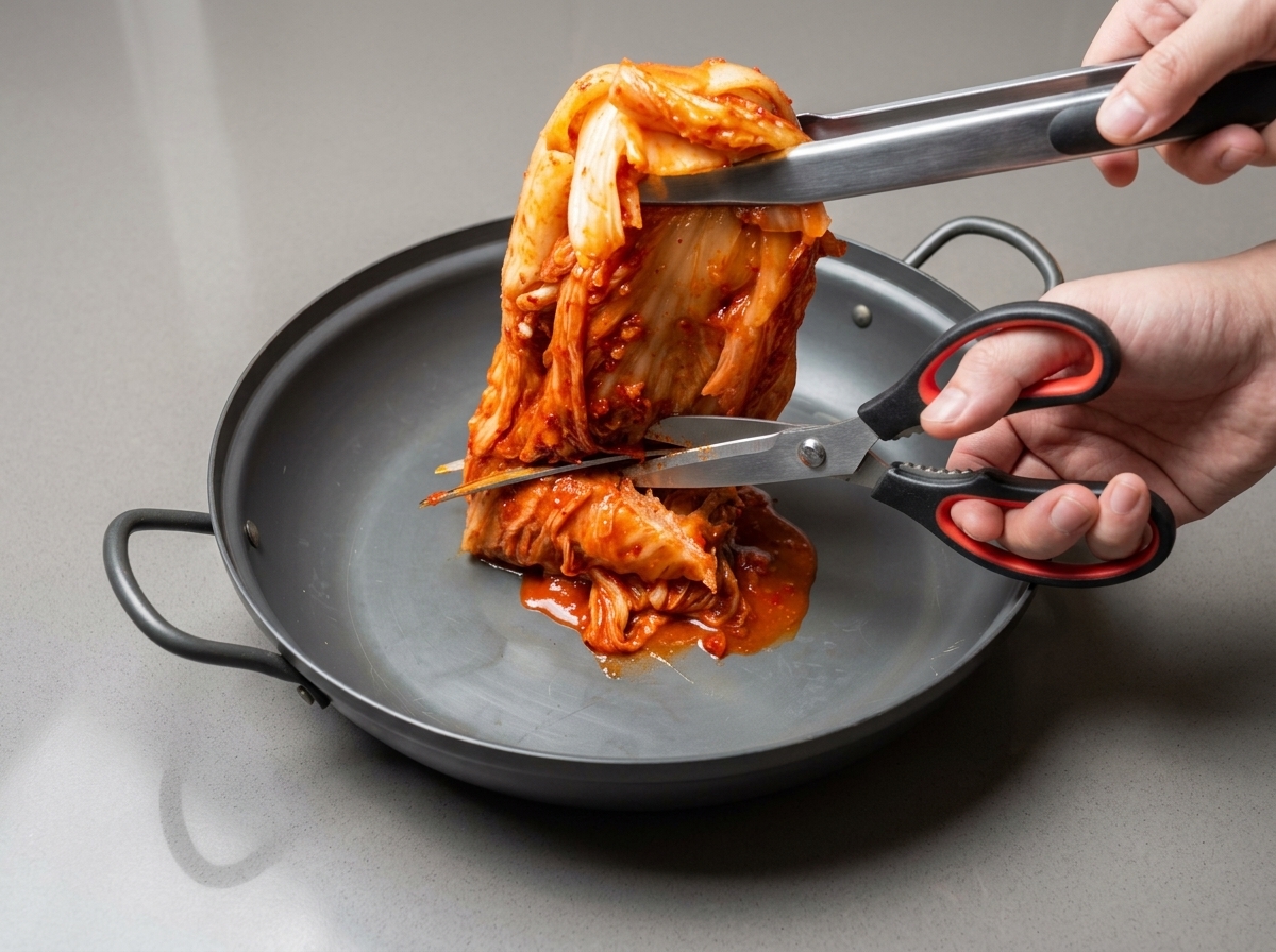 Tijeras de cocina cortando kimchi entero en trozos más pequeños dentro de una olla ancha.