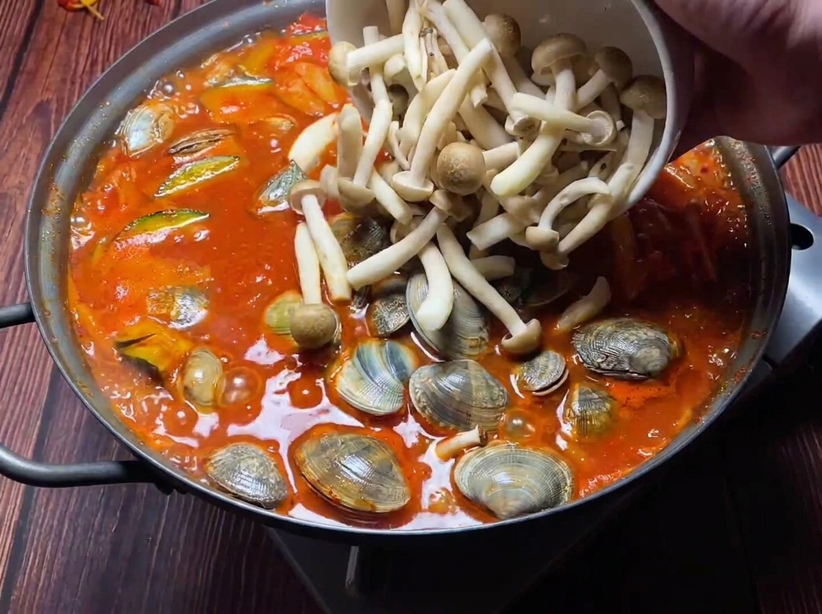 Almejas frescas y setas variadas añadiéndose a un estofado rojo de kimchi burbujeante.