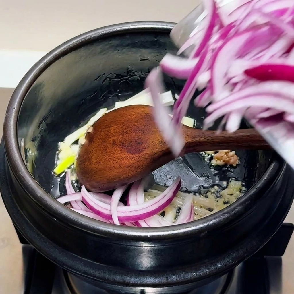 Cebollas rojas en rodajas, ajo picado y partes blancas de cebollino siendo salteadas en una olla negra con una cuchara de madera.
