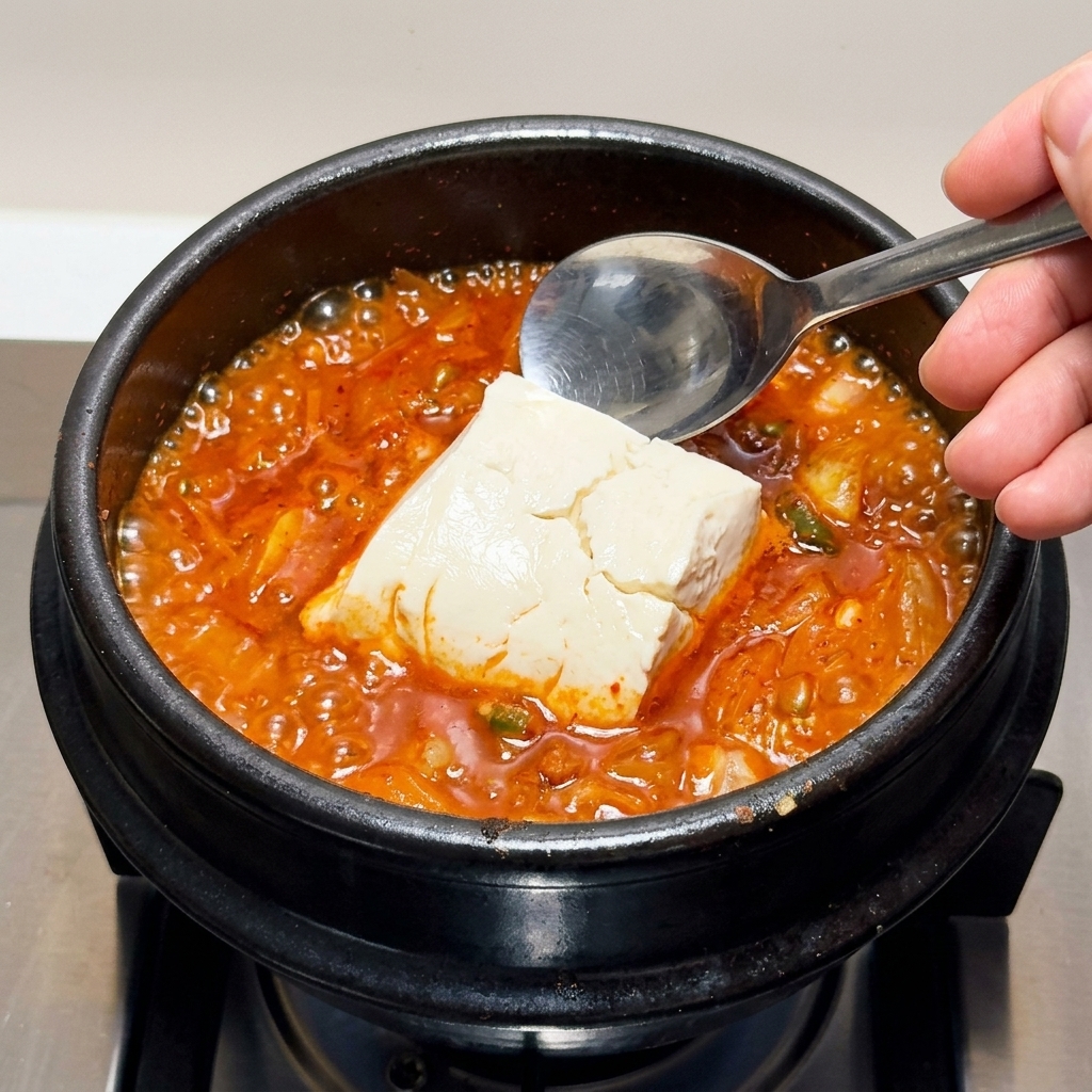 Grandes trozos de tofu sedoso siendo colocados con cuidado en el guiso de kimchi hirviendo.