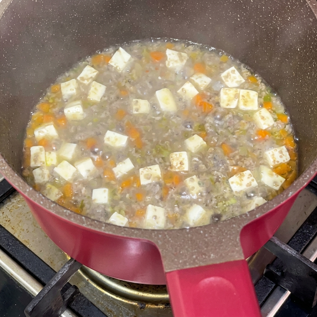 Salsa de soja siendo añadida a una mezcla de carne, tofu y zanahorias cocinándose a fuego lento.
