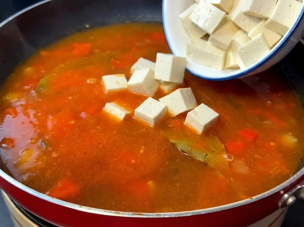 Cubos de tofu sedoso y ternera salteada siendo añadidos a la base de sopa de tomate hirviendo.