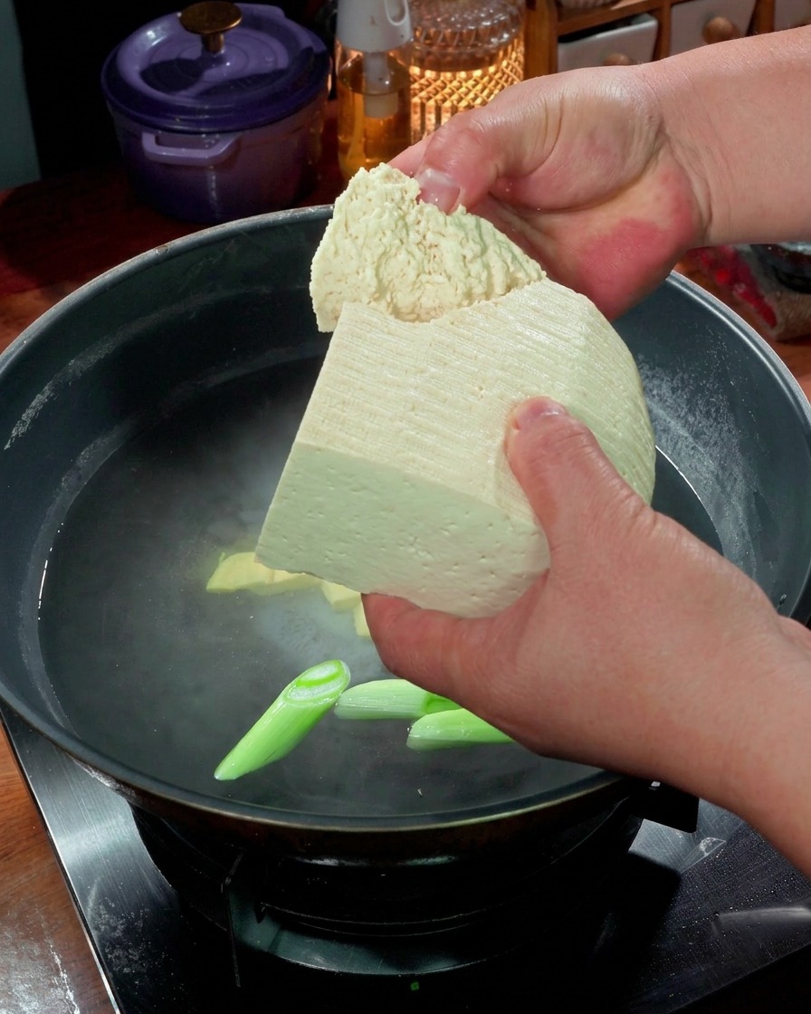 Manos rompiendo trozos grandes de tofu firme en una olla de agua hirviendo con cebolletas.