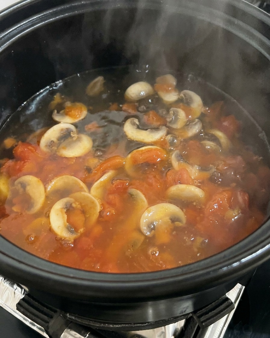 Agua hirviendo siendo vertida en una olla llena de tomates y champiñones salteados, creando vapor.