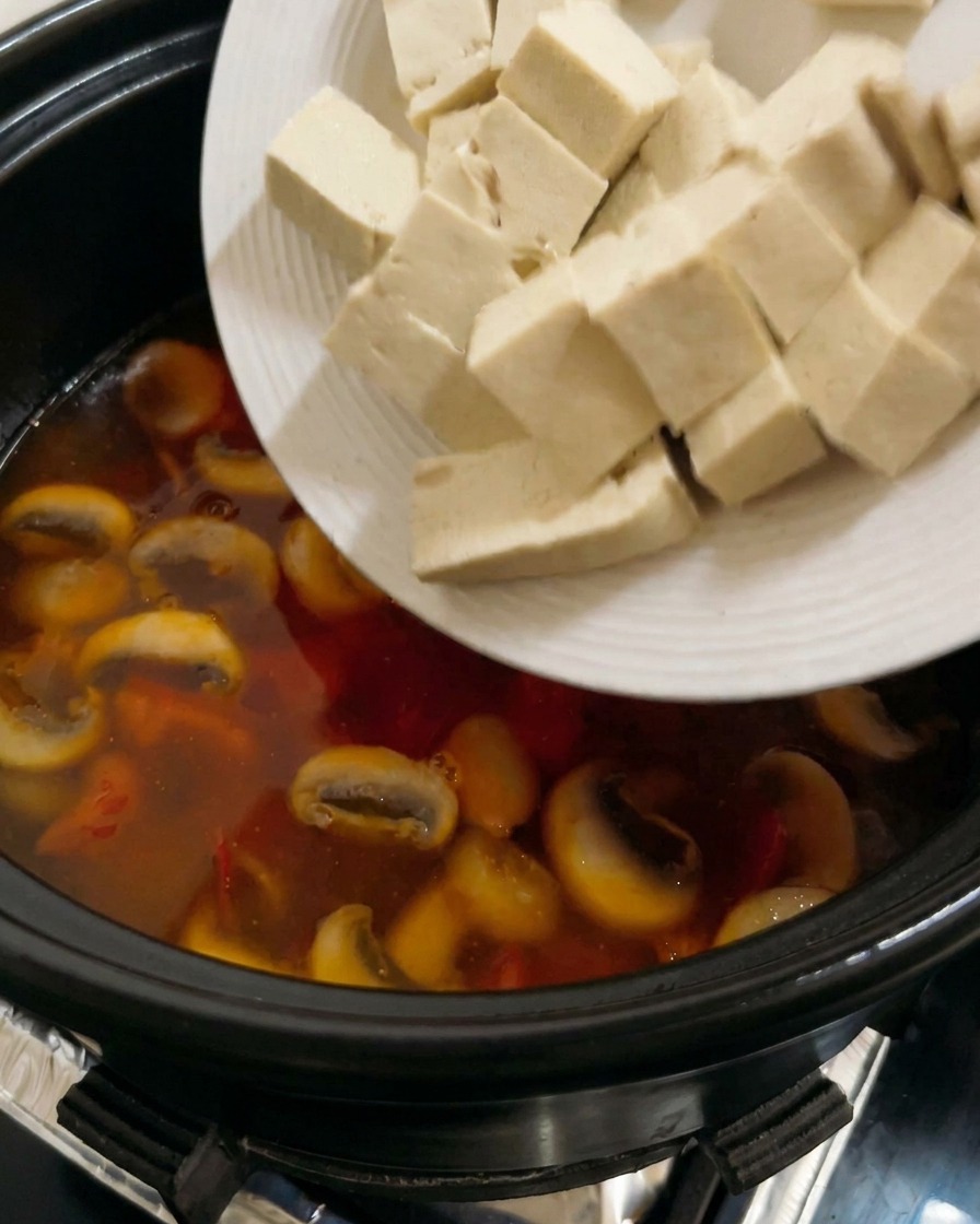 Cubos de tofu blanco firme siendo añadidos a una sopa de tomate y champiñones burbujeante.