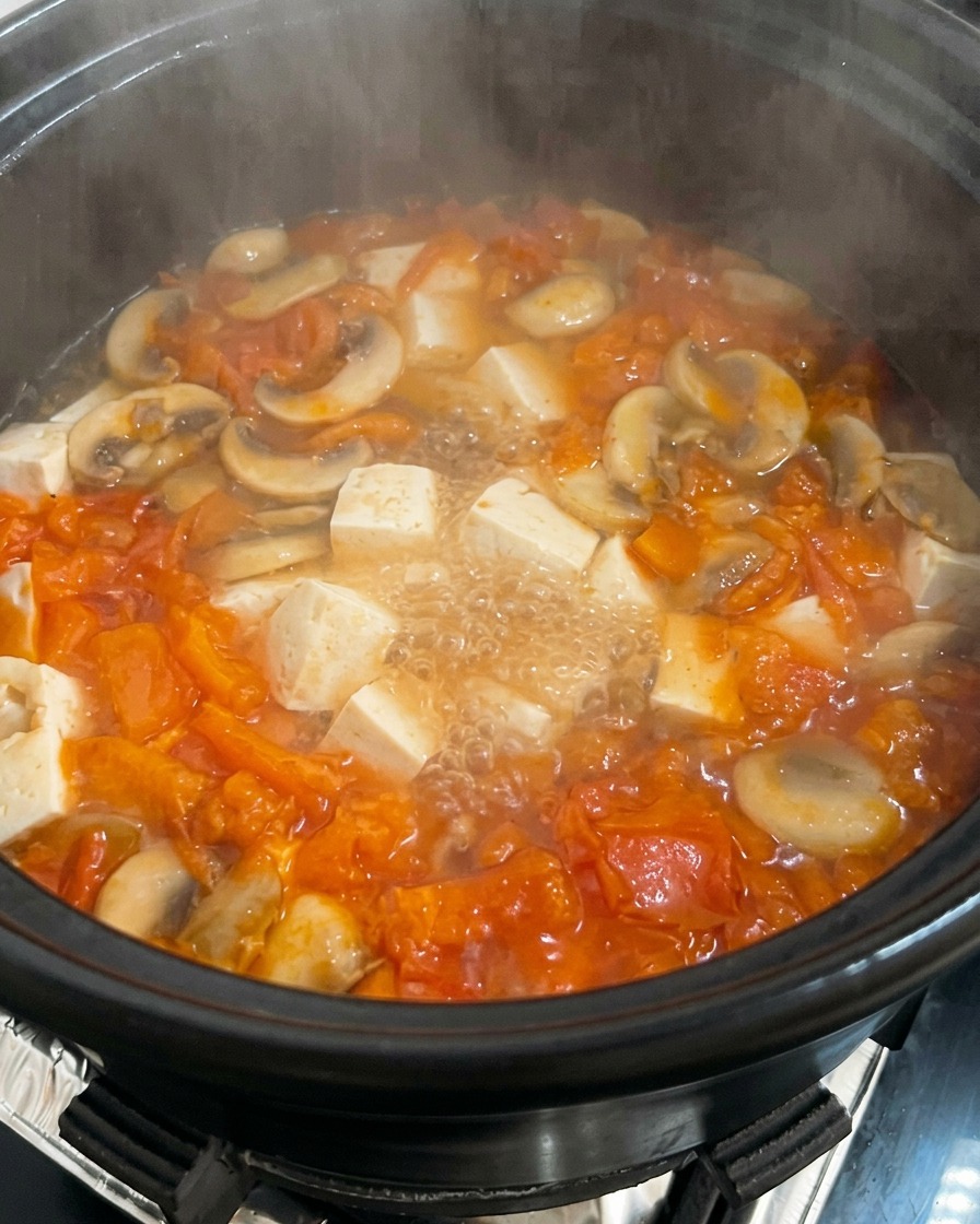 Una olla de sopa de tomate y champiñones a fuego lento con cubos de tofu, teniendo huevo batido siendo vertido en ella.