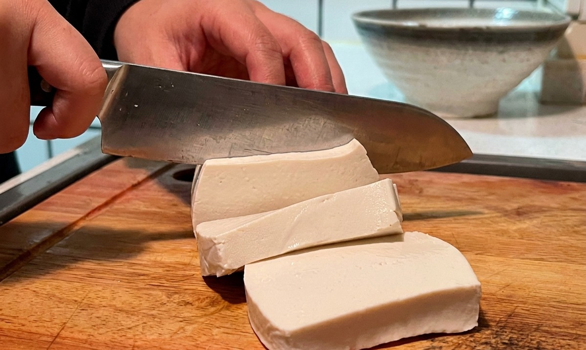 Un bloque de tofu blanco siendo cortado en cubos pequeños sobre una tabla de madera.