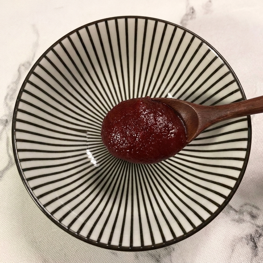 Una cuchara de madera añadiendo una cucharada de pasta de chile coreano roja en un tazón con patrones de rayas.