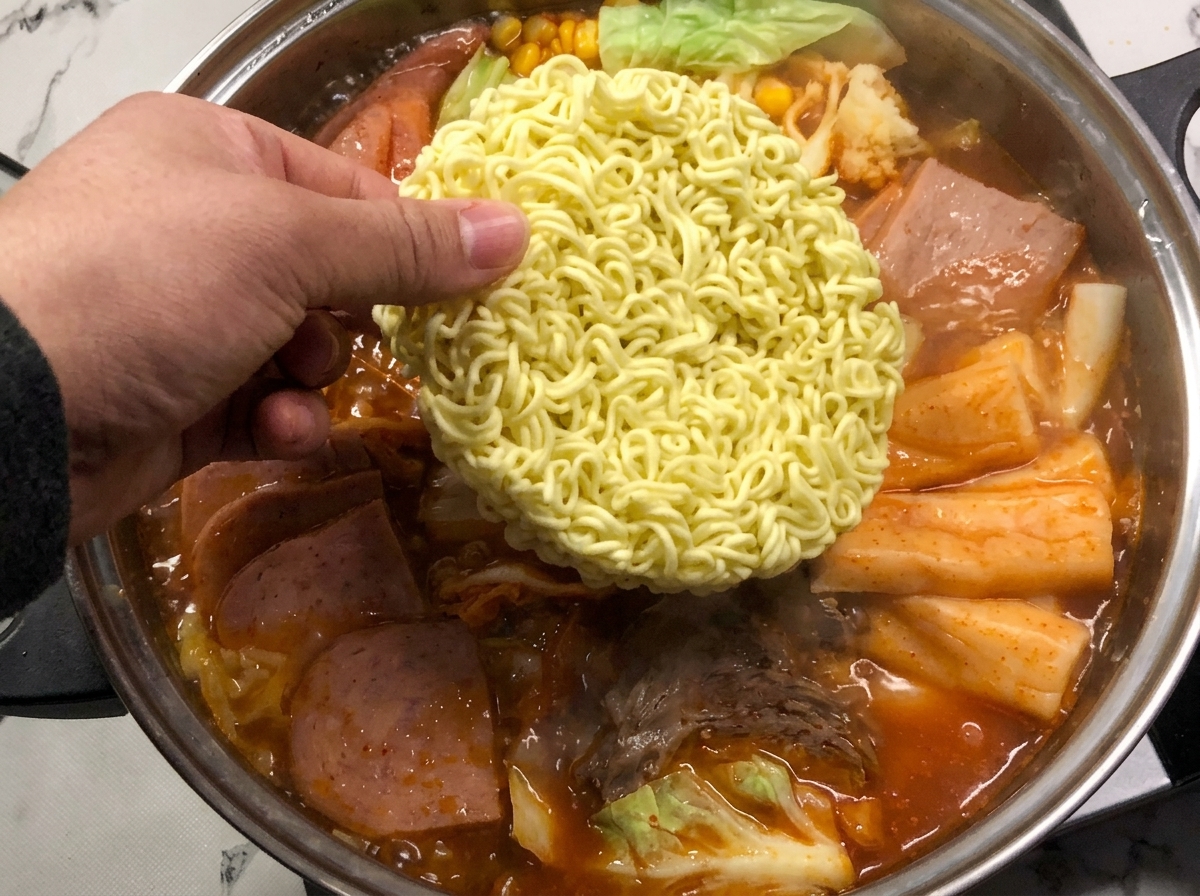 Un bloque de fideos instantáneos siendo bajado al vibrante caldo rojo burbujeante del estofado.