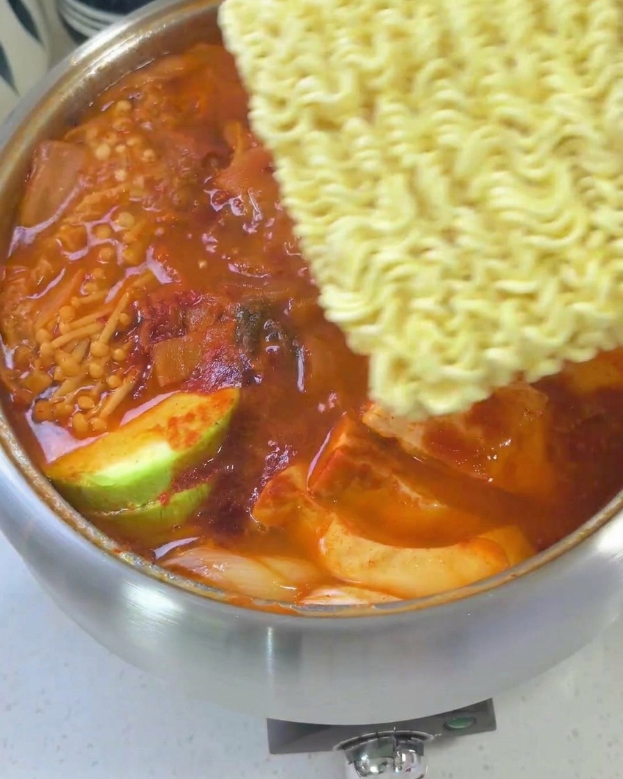Un estofado coreano casi terminado cubierto con fideos ramen, lonchas de queso amarillo y un huevo, listo para la cocción final.