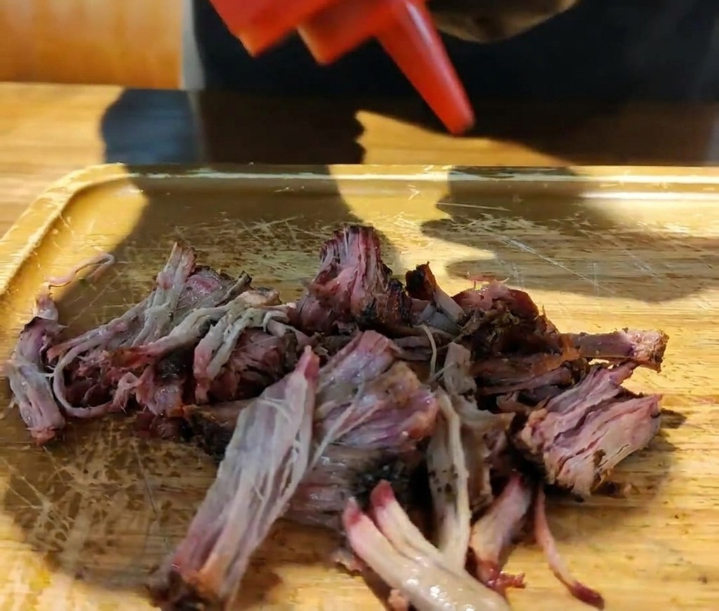Un bote rojo vertiendo salsa barbacoa espesa sobre una pila de cerdo ahumado desmenuzado en una tabla de madera.