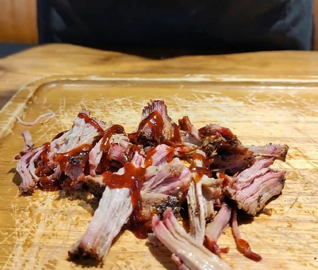 Una mano añadiendo una mezcla de repollo morado y blanco picado sobre el cerdo desmenuzado con salsa en una tabla de madera.