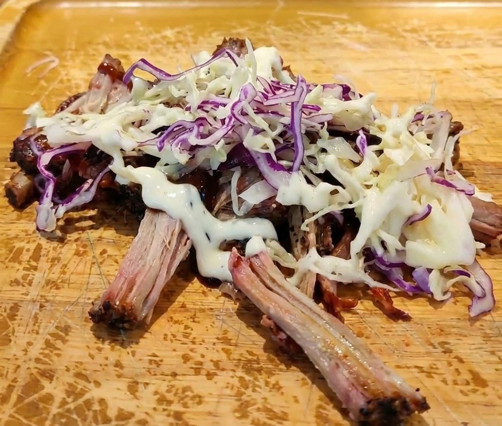 Cerdo desmenuzado con salsa BBQ cubierto con una montaña de ensalada de col fresca y un chorro de aderezo blanco cremoso.