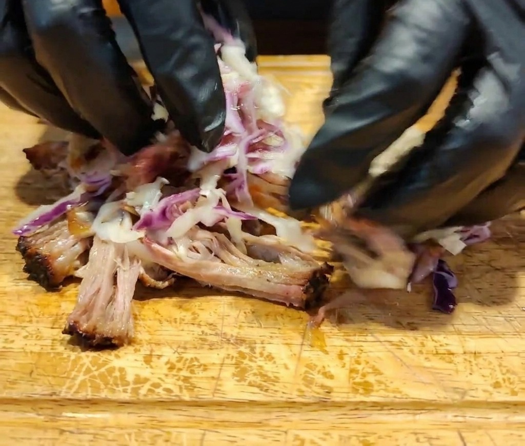 Manos con guantes mezclando carne desmenuzada, salsa oscura y ensalada de col colorida sobre una superficie de madera.