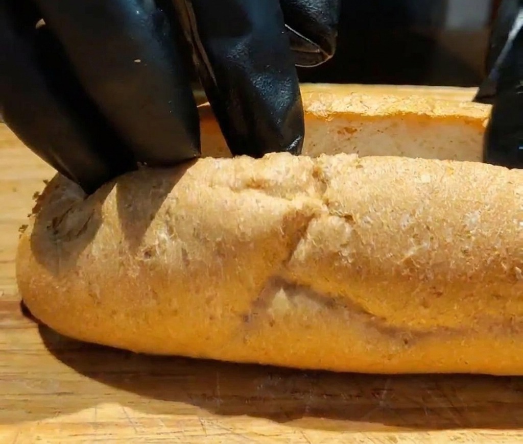 Primer plano de manos con guantes negros abriendo una baguette integral cortada en una tabla de madera para crear espacio.
