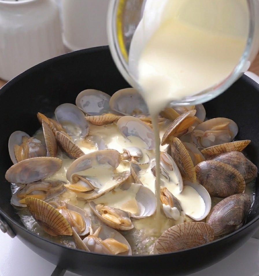 Crema de leche espesa vertiéndose desde una jarra medidora sobre las almejas abiertas.