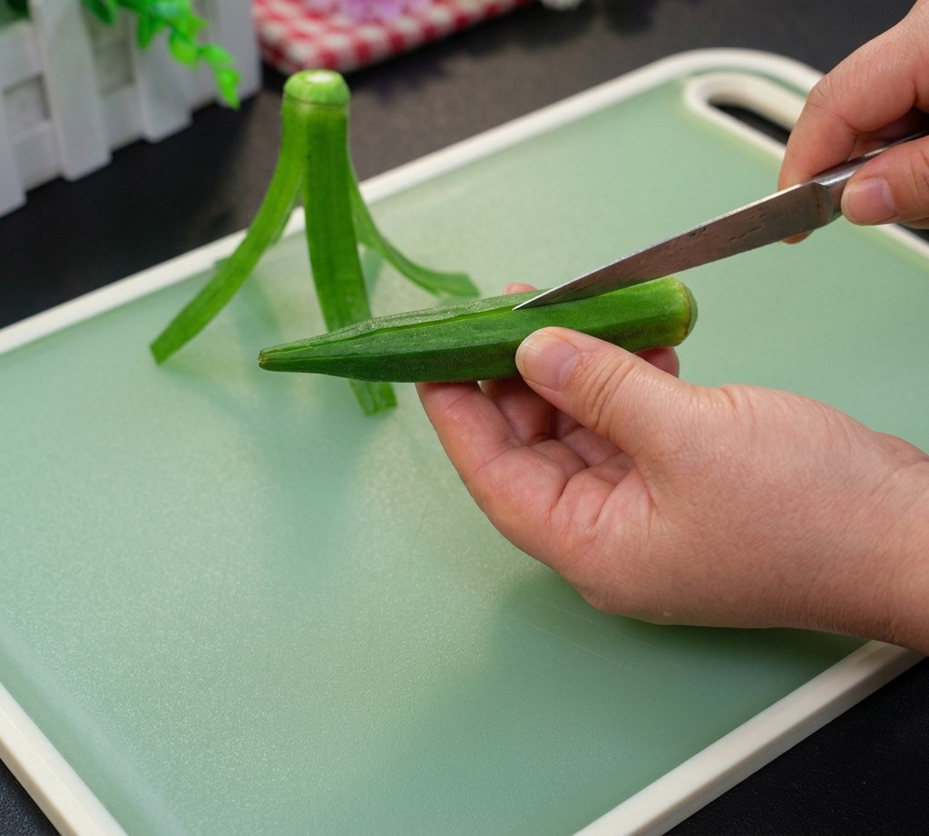 Una mano cortando el borde superior de una okra verde sobre una tabla de cortar.
