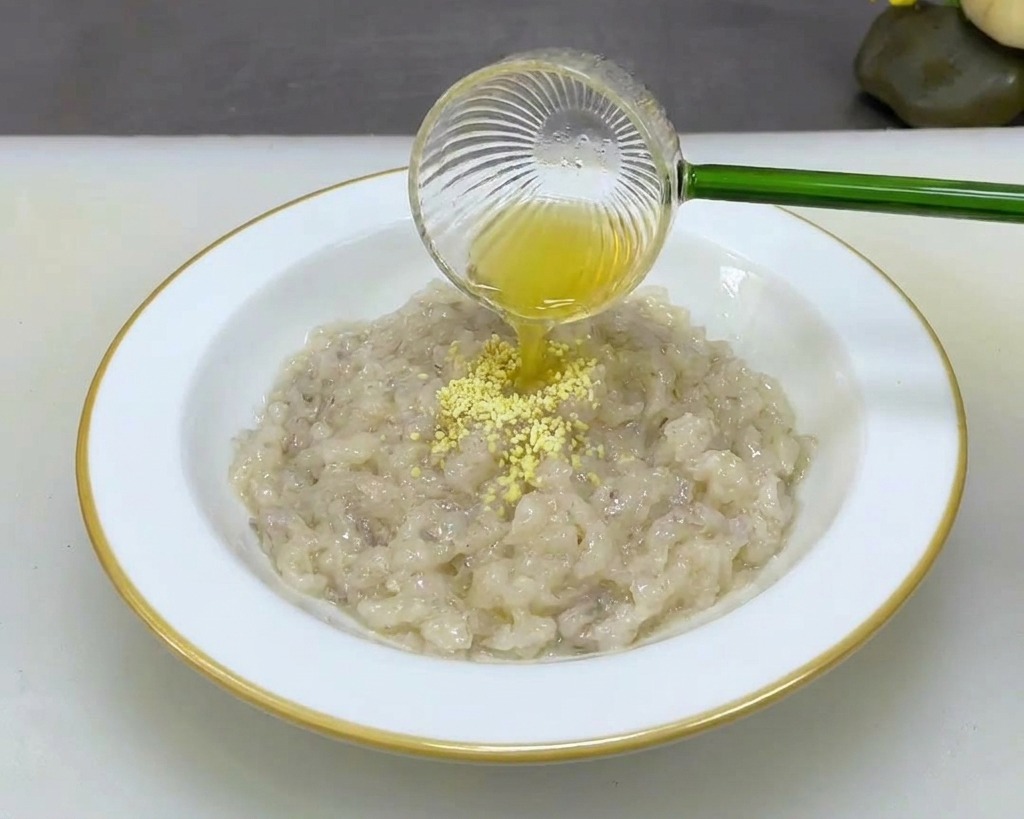 Persona vertiendo condimento en polvo desde una pequeña cuchara de cristal a un bol blanco con pasta de camarón espesa.