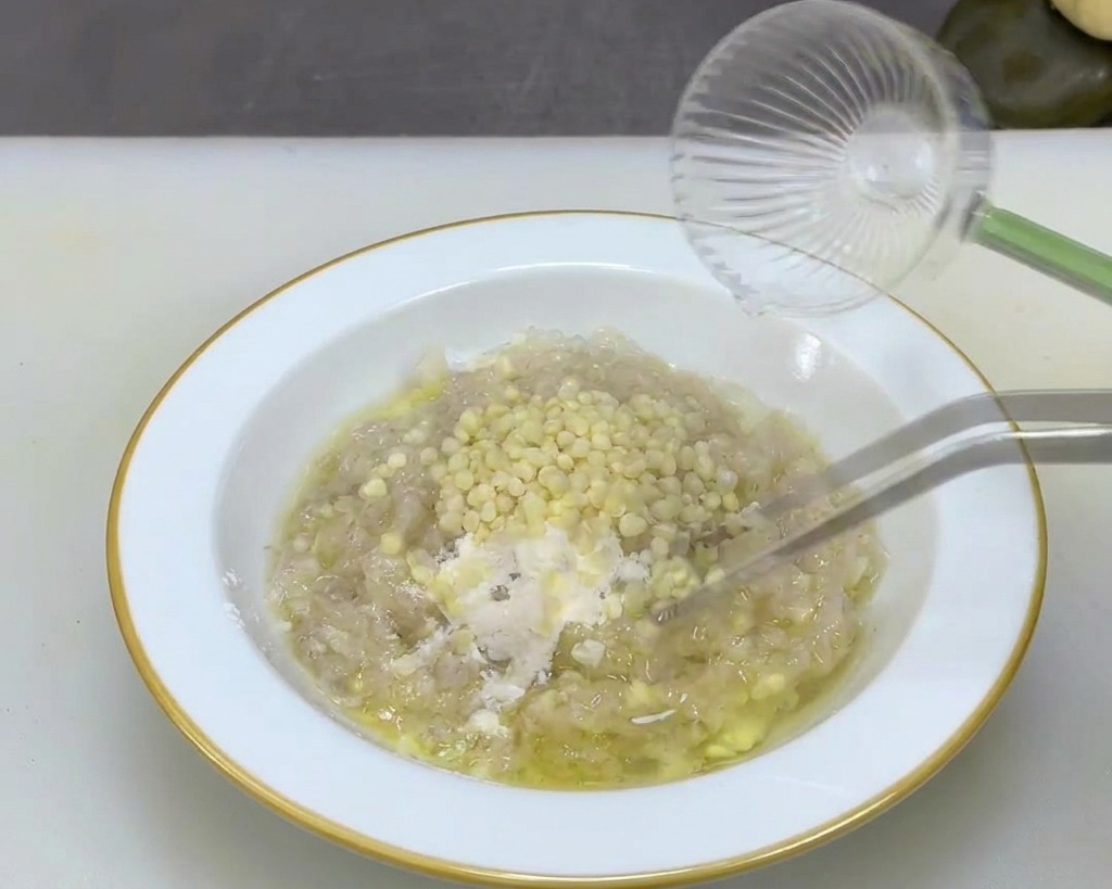 Pinzas metálicas mezclando pequeñas semillas de okra color crema en una pasta de camarón espesa en un bol blanco.