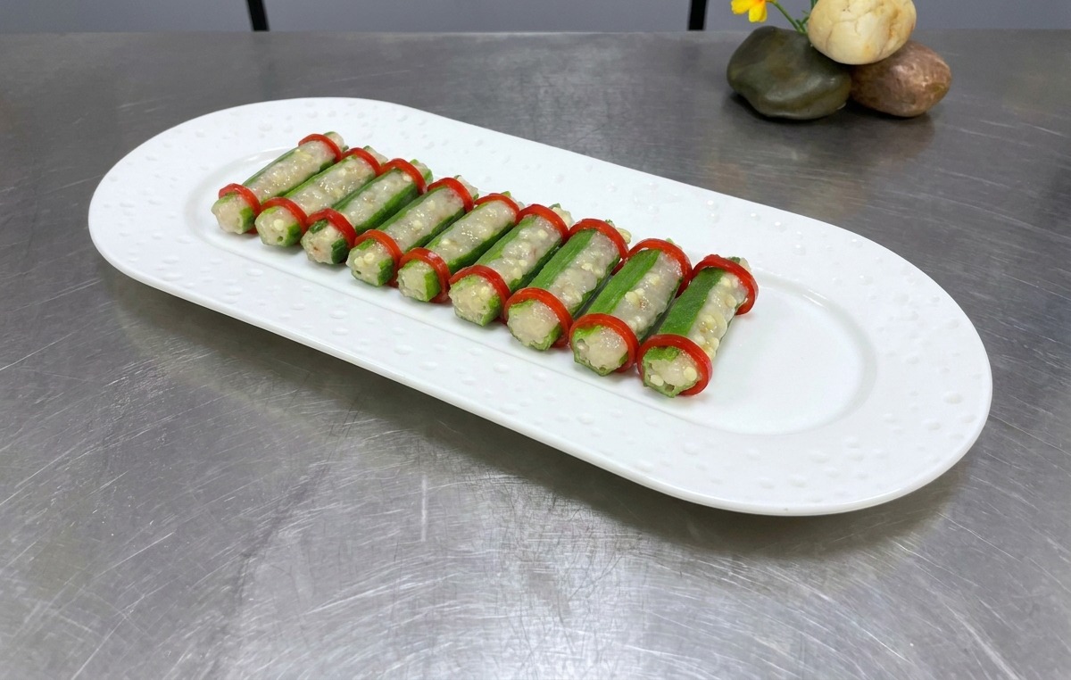Dos filas ordenadas de okra rellena con aros de chile rojo en un plato ovalado blanco, listo para cocinar al vapor.