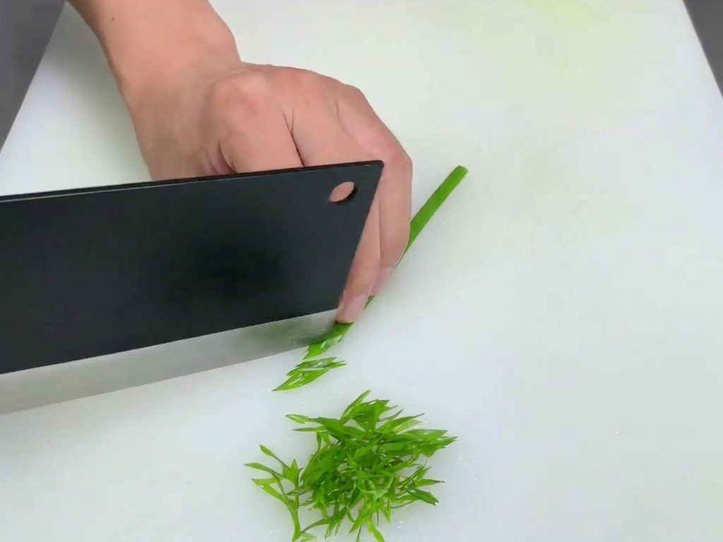Persona usando un cuchillo grande para picar finamente tallos de cebolleta sobre una tabla de cortar blanca.