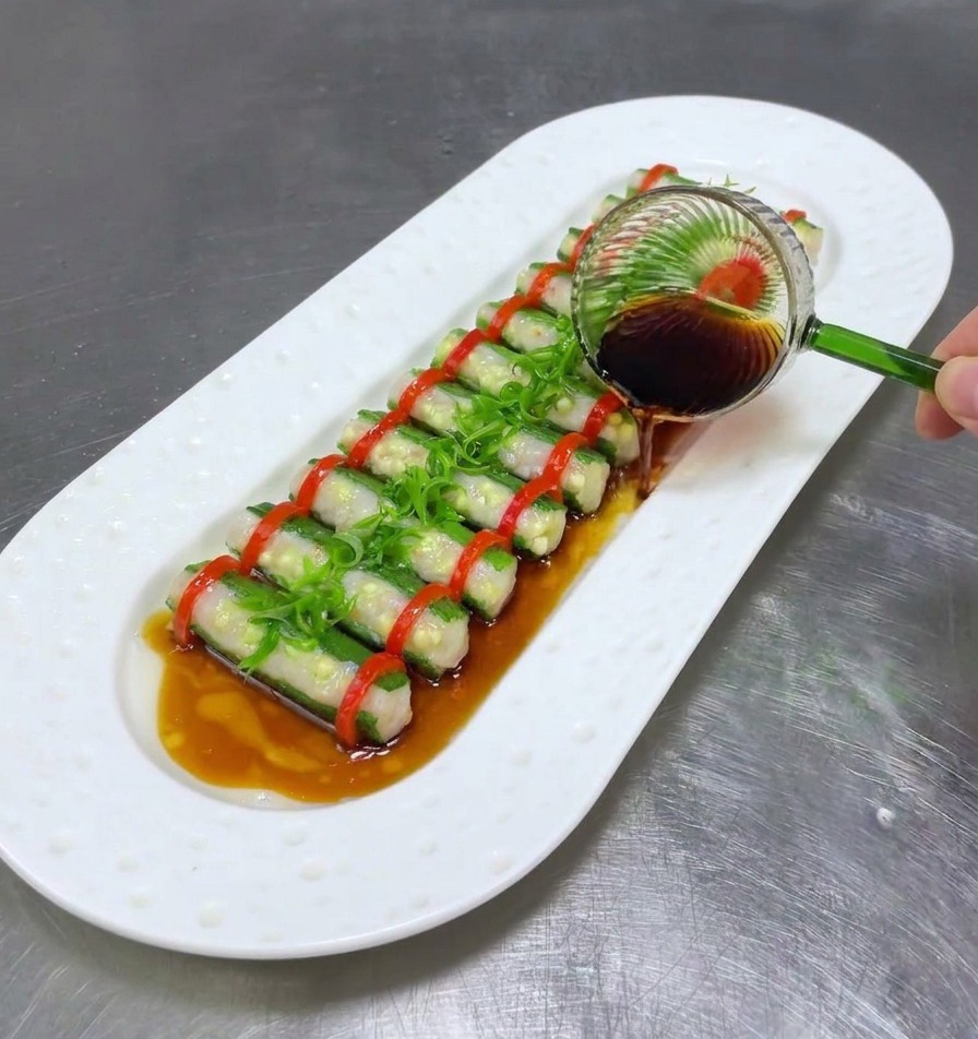 Cucharón de cristal rociando salsa de soja oscura sobre la okra rellena al vapor y las cebolletas en un plato blanco.