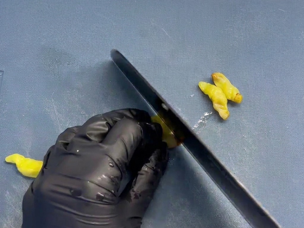 Un chef usando un cuchillo negro para picar finamente pequeños chiles amarillos encurtidos sobre una tabla de cortar azul.