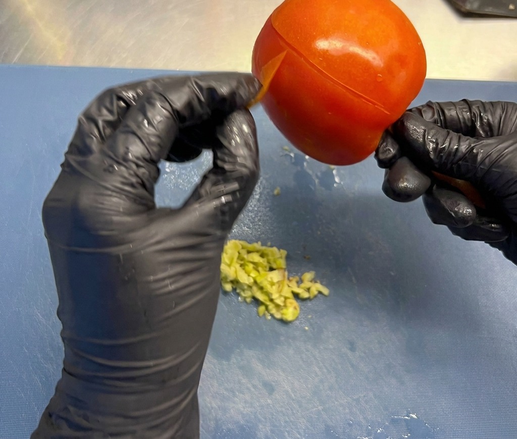 Manos con guantes negros quitando la fina piel exterior de un tomate rojo grande y maduro sobre una tabla de preparación.