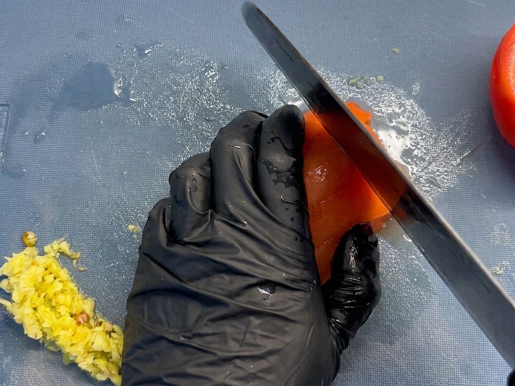 Primer plano de un chef cortando un tomate rojo pelado en trozos uniformes sobre una tabla de cortar azul.