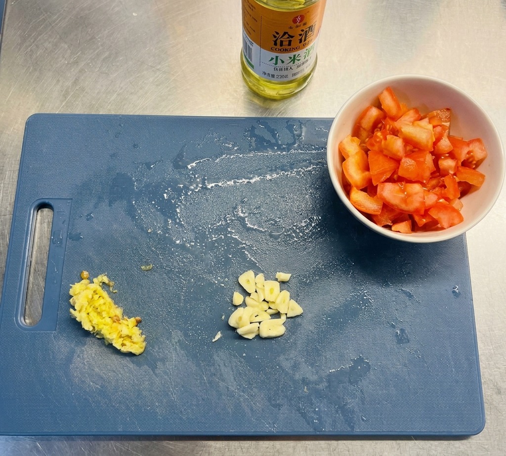 Una tabla de cortar azul que muestra montones de ajo laminado, chiles amarillos picados y tomates troceados junto a una botella de aceite.