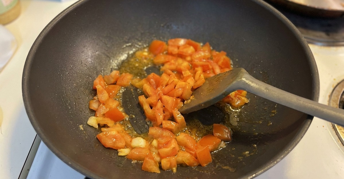 Trozos de tomate picado siendo salteados con ajo en un wok oscuro, con jugos comenzando a burbujear en el fondo.