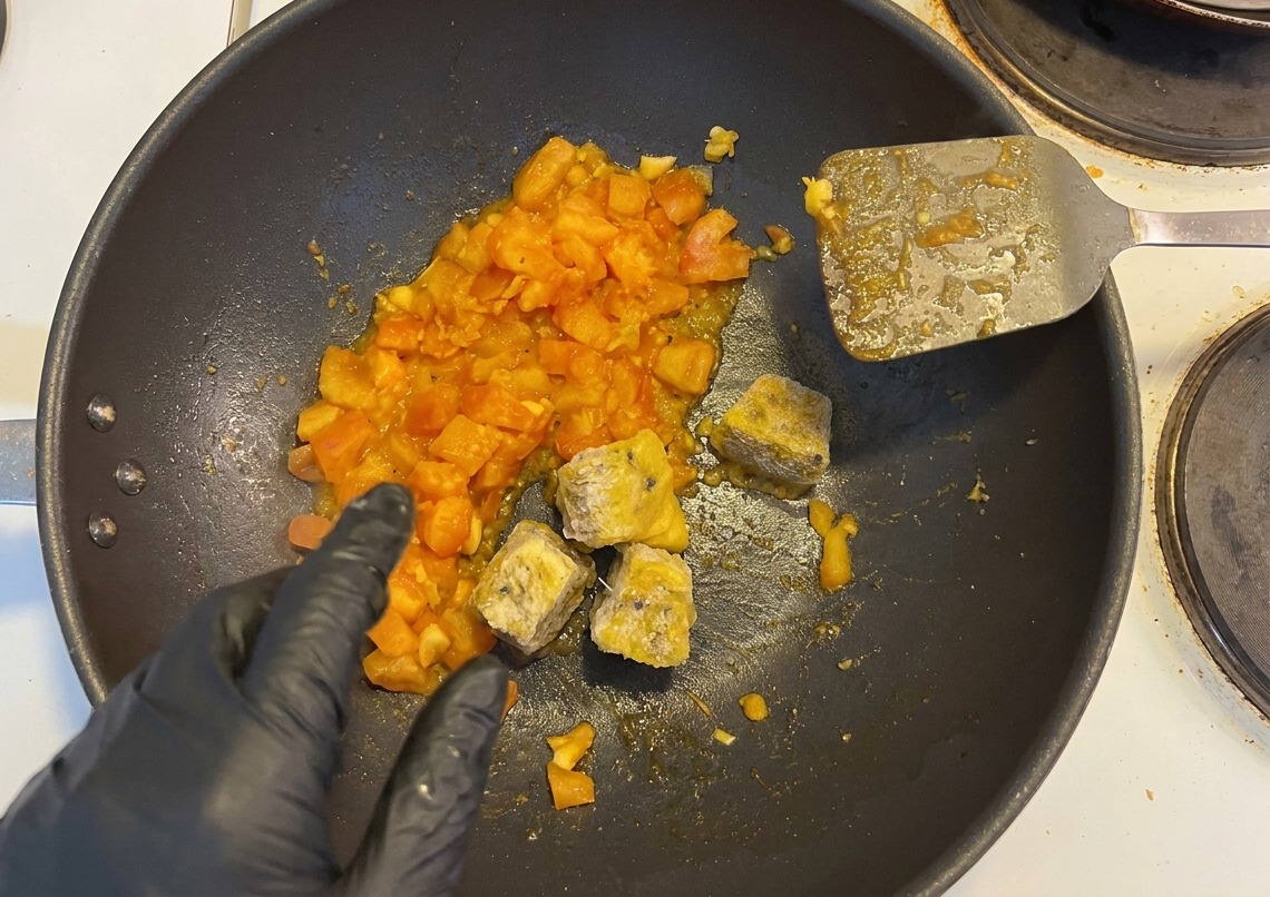Cubos congelados de pulpa de maracuyá y chiles encurtidos picados siendo añadidos a una sartén con tomates salteados por una mano con guante negro.