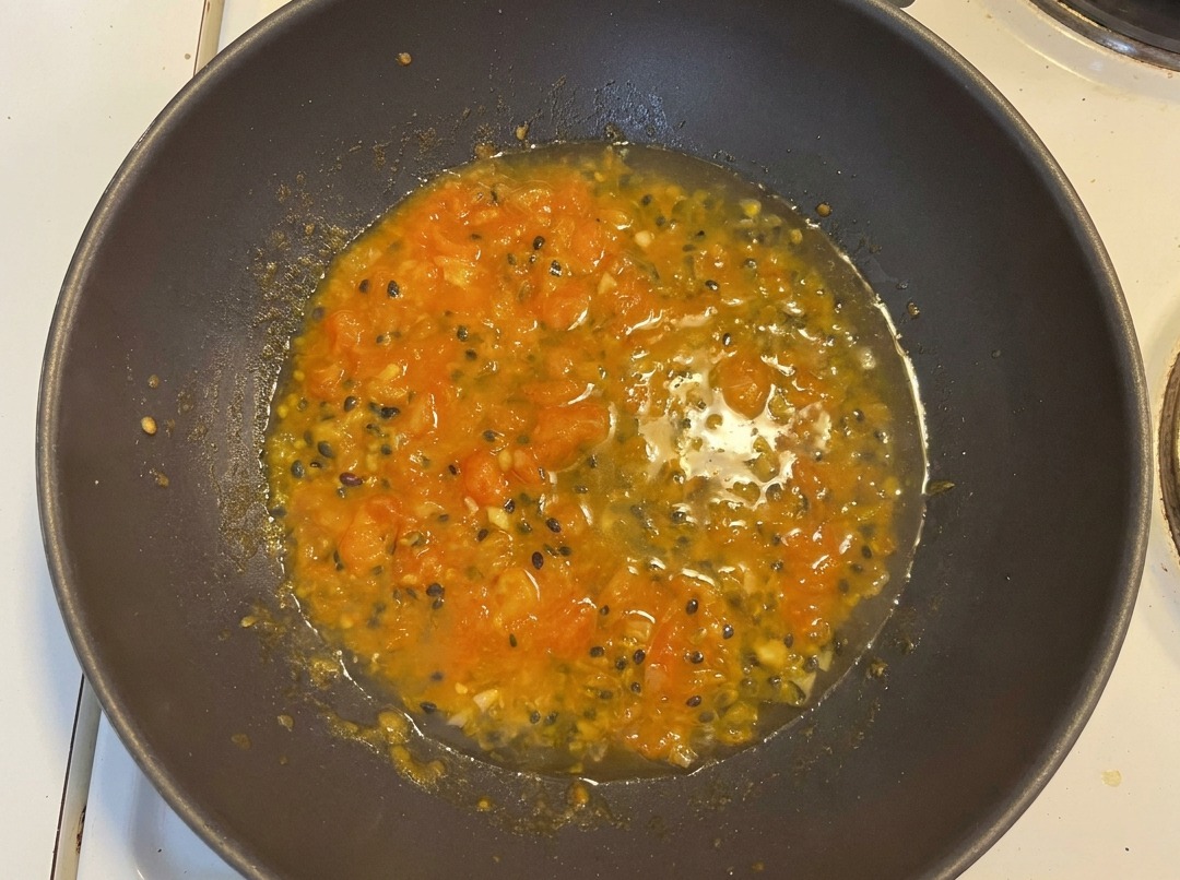 Agua añadida a la mezcla de tomate y maracuyá en un wok oscuro, creando una base de sopa naranja vibrante con semillas de maracuyá visibles.