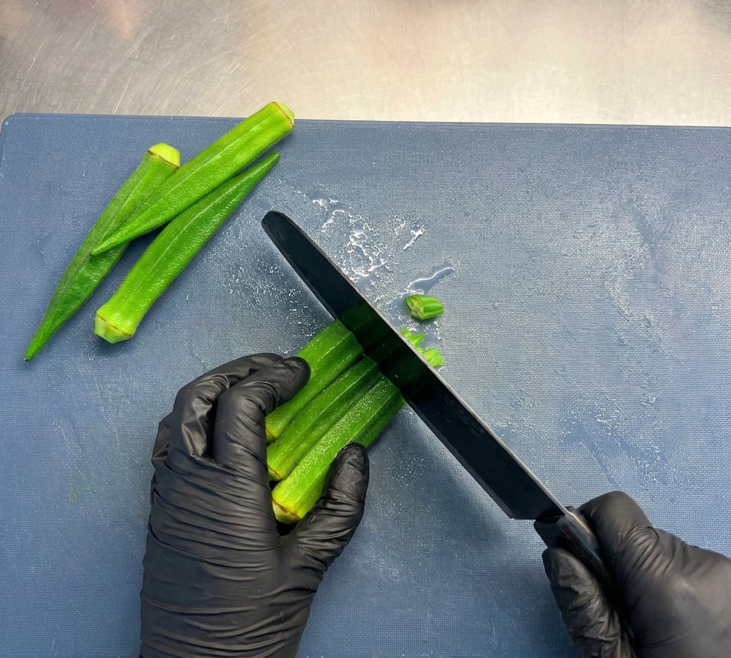 Una persona con guantes negros usando un cuchillo de chef para cortar tallos de quingombó verde brillante en pequeñas rodajas circulares sobre una tabla de cortar azul.