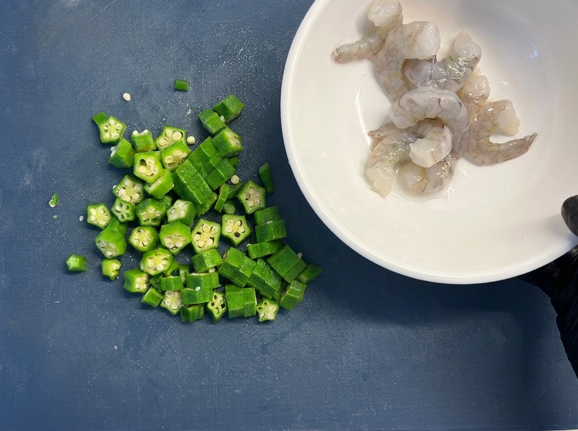Un tazón blanco lleno de camarones crudos y limpios sostenido junto a un montón de rodajas de quingombó verde en una tabla de cortar azul.