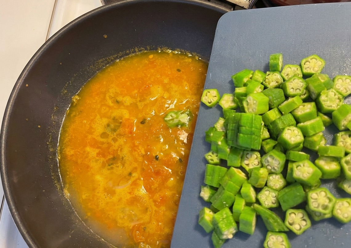 Rodajas de quingombó recién cortadas deslizándose desde una tabla de cortar azul hacia una sopa naranja burbujeante en un wok oscuro.