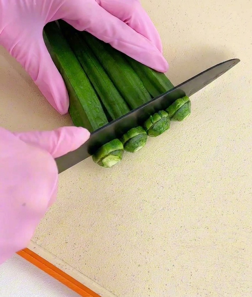 Manos con guantes rosas usando un cuchillo negro para cortar los extremos del tallo de un manojo de okra verde.