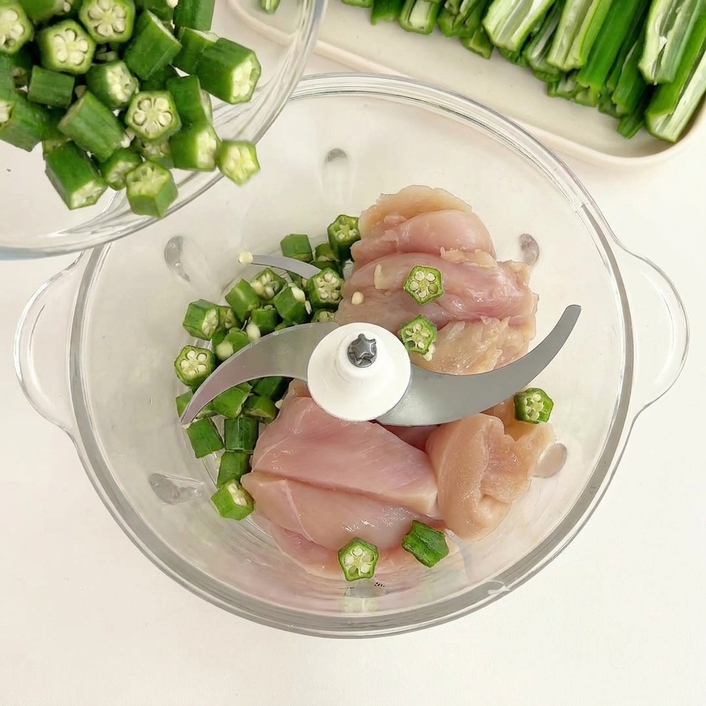 Semillas de okra verde cayendo desde un bol pequeño sobre trozos de pollo crudo en un procesador de vidrio.