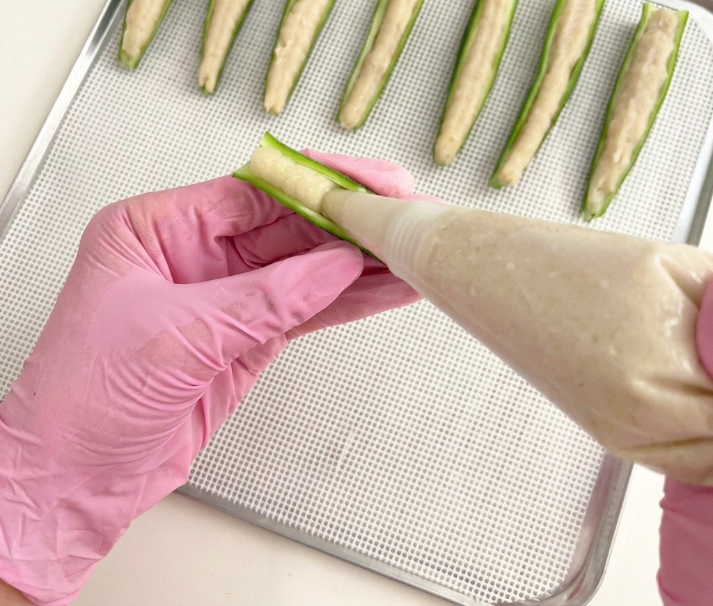 Una persona con guantes rosas usando una manga pastelera para rellenar mitades de okra verde con pasta de pollo sobre una rejilla metálica.