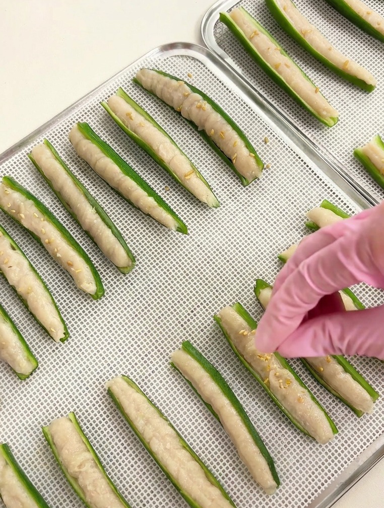 Primer plano de una mano espolvoreando semillas de sésamo blanco sobre filas de okra rellena en una bandeja plateada.