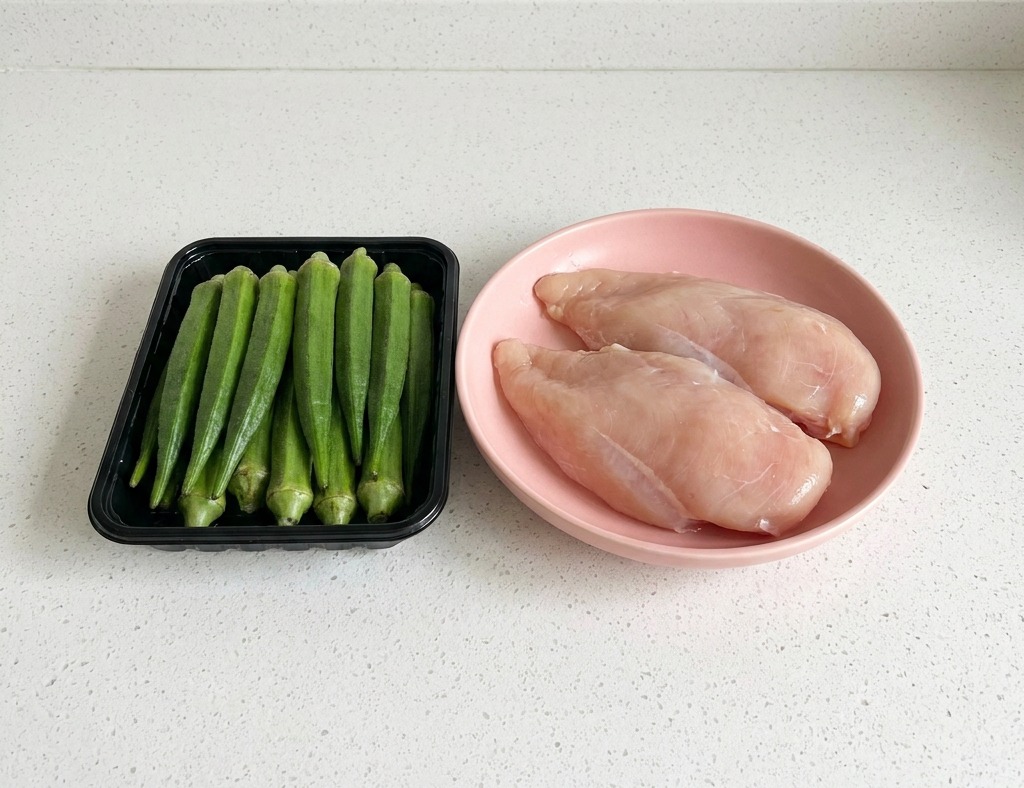 Una bandeja negra con okra verde fresca junto a un plato con dos filetes de pechuga de pollo cruda sobre una encimera blanca.
